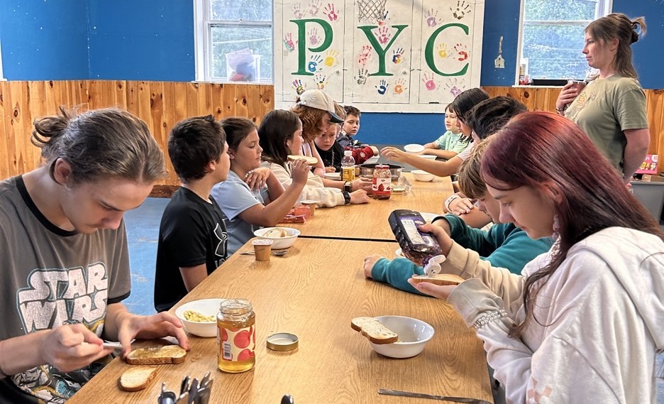 Children sitting at a table, preparing sandwiches, overseen by a counselor. The letters "PYC" are clear in the background.