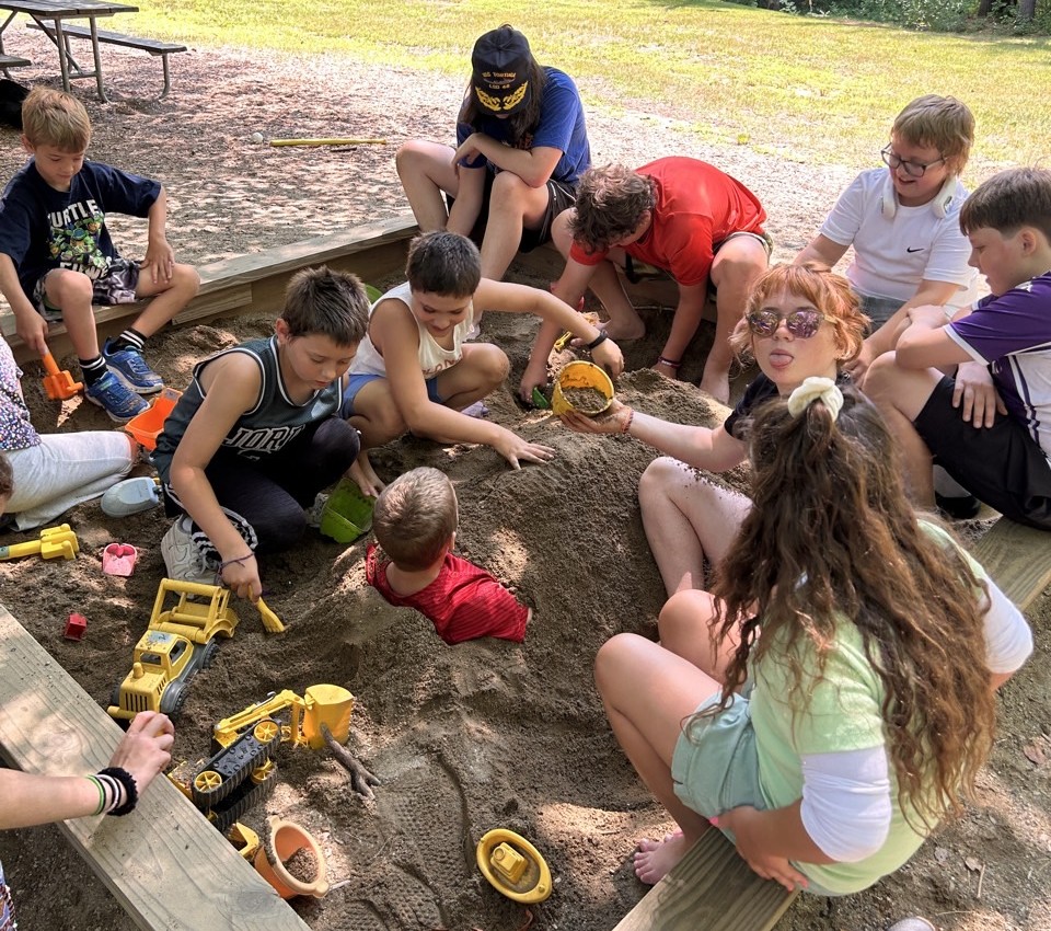 Children playing in the sandbox, covering one child up to the shoulders in sand.