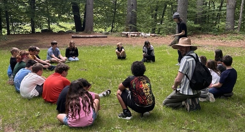 Children sitting in a circle in the green grass at the park. A counselor joins them, smiling.
